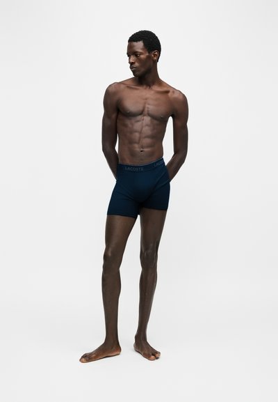 Homme musclé debout pieds nus portant un boxer Lacoste bleu foncé, regardant vers sa gauche sur un fond clair uni.