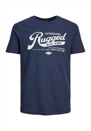 Mężczyzna w granatowym T-shircie "Jack & Jones Rugged Blue Jeans" i jasnoniebieskich podartych dżinsach na jednolitym tle.