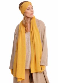 Écharpe en tricot jaune associée à un bandeau assorti, tous deux dans une texture douce. La tenue comprend un manteau beige et des gants, créant un look cohérent.