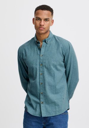 Overhemd van teal stof met knoopsluiting, voorzien van een puntige kraag, lange mouwen en een rechte zoom. Zes bruine knopen versieren de voorkant.