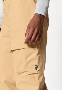 Calças cargo bege feitas de tecido leve, com um grande bolso lateral e um patch de logotipo preto na parte superior da perna.