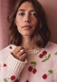 Maglione lavorato a maglia in rosa tenue con collo a coste. Presenta un motivo di ciliegie e foglie verdi. Il modello mostra un anello d'argento sulla mano.