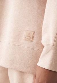 Gros plan sur le bord-côte et l'ourlet d'un sweat-shirt beige avec un logo carré cousu représentant la silhouette d'un joueur de basketball et une signature.