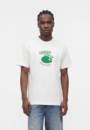 Jeune homme aux cheveux tressés portant un t-shirt blanc avec un logo grenouille verte et le texte « CARHARTT WIP please don’t snitch » et un jean bleu.
