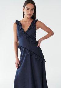 Laivastonsininen mekko syvällä V-päällä, röyhelöreunoilla ja epäsymmetrisellä helmalla. Valmistettu teksturoituneesta kankaasta, jossa on muotoiltu yläosa ja kunnollinen hame.