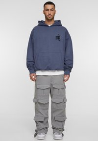 Übergroßer navy Hoodie mit schwarzem Logo und Fronttasche, kombiniert mit grauen Cargo-Hosen, die mehrere Taschen und eine lockere Passform bieten.