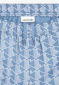 Μπλε μαγιό σορτς με γεωμετρικό λευκό σχέδιο. Διαθέτει ελαστική μέση με κορδόνι και ετικέτα με το λογότυπο της Lacoste.