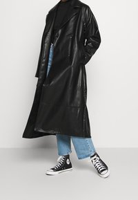 Trench-coat en cuir noir avec un design croisé, de larges revers et une finition brillante, associé à un jean court bleu clair et des baskets noires.