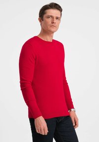 Pull en tricot rouge avec des manches longues et un col rond, fabriqué à partir d'un tissu au toucher lisse, associé à un jean foncé. Design simple, sans motifs.
