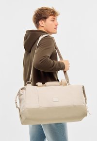 Beige Reisetasche mit einem helleren Oberteil und einem dunkleren Boden, hergestellt aus strapazierfähigem Material. Verfügt über Tragegriffe und einen verstellbaren Schultergurt.