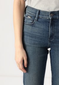 Mørkeblå denim jeans med høy midje, rette ben, med en frontlomme og merke. Hånd synlig på siden.