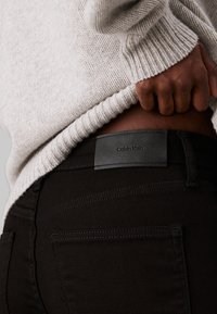 Melns džinsu audums ar ādas Calvin Klein birku aizmugurē. Gaiši pelēks adīts džemperis ir daļēji redzams.