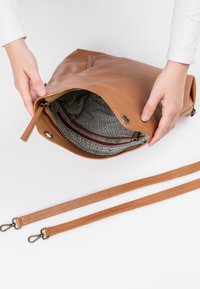 Braune Ledertasche mit einem Reißverschluss, die ein gemustertes Innenfutter zeigt. Mit zwei langen Lederträgern mit Metallclips und Branding-Detail.