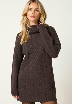 Femme blonde portant une longue robe-pull à torsades de couleur marron foncé avec un col roulé côtelé haut.