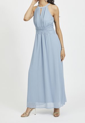 Femme portant une robe longue bleu pâle sans manches avec une taille en dentelle et un col en forme de goutte, associée à des sandales à talons hauts dorées.