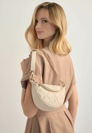 Kobieta o blond włosach, ubrana w beżową sukienkę, trzyma na ramieniu pikowaną kremową torebkę na złotym łańcuszku z zamkiem, patrząc za siebie.