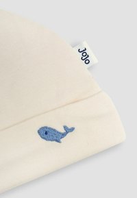 Gorro de algodón color crema con un diseño de ballena bordado en azul y una pequeña etiqueta tejida que dice "Jojo". Textura suave y lisa.