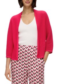 Rosa geripptes Strickjacke mit offenem Vorderteil, Dreiviertelärmel, kombiniert mit einem weißen V-Ausschnitt-Oberteil und gemusterten Hosen mit rosa und schwarzen Pfeilen.