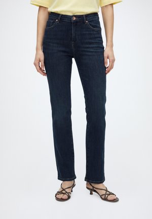 Person iført mørkeblå jeans med lige ben, gul kortærmet top og brune sandaler med hæl og remme, stående mod en ensfarvet baggrund.