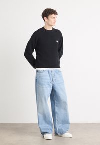 Zwarte geribbelde trui met een klein logo-detail, gecombineerd met lichtblauwe wijde jeans en witte sneakers op een neutrale achtergrond.
