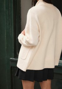 Roomkleurige oversized cardigan gemaakt van zachte stof, met geribbelde boorden en zoom, en een zak aan de zijkant.