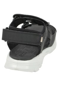 Svart atletisk sandal med justerbara remmar, en texturerad svart innersula och en vit gummisul, med en logotyp på sidan.