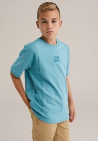 Turkiozine trumpa rankovė t-shirt su juostos tipo apykakle ir subtiliu užrašu "BLUE RIDGE" atitinkama spalva. Minkšta medvilnės mišinio medžiaga, laisvas siluetas.