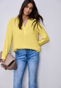 Femme aux longs cheveux bruns portant un chemisier jaune et un jean bleu, tenant un sac à main beige, debout devant un mur clair uni.