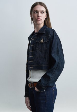 Mujer joven con chaqueta y jeans de mezclilla oscuros, con la mano en el bolsillo, mirando de lado sobre un fondo liso de color gris claro.