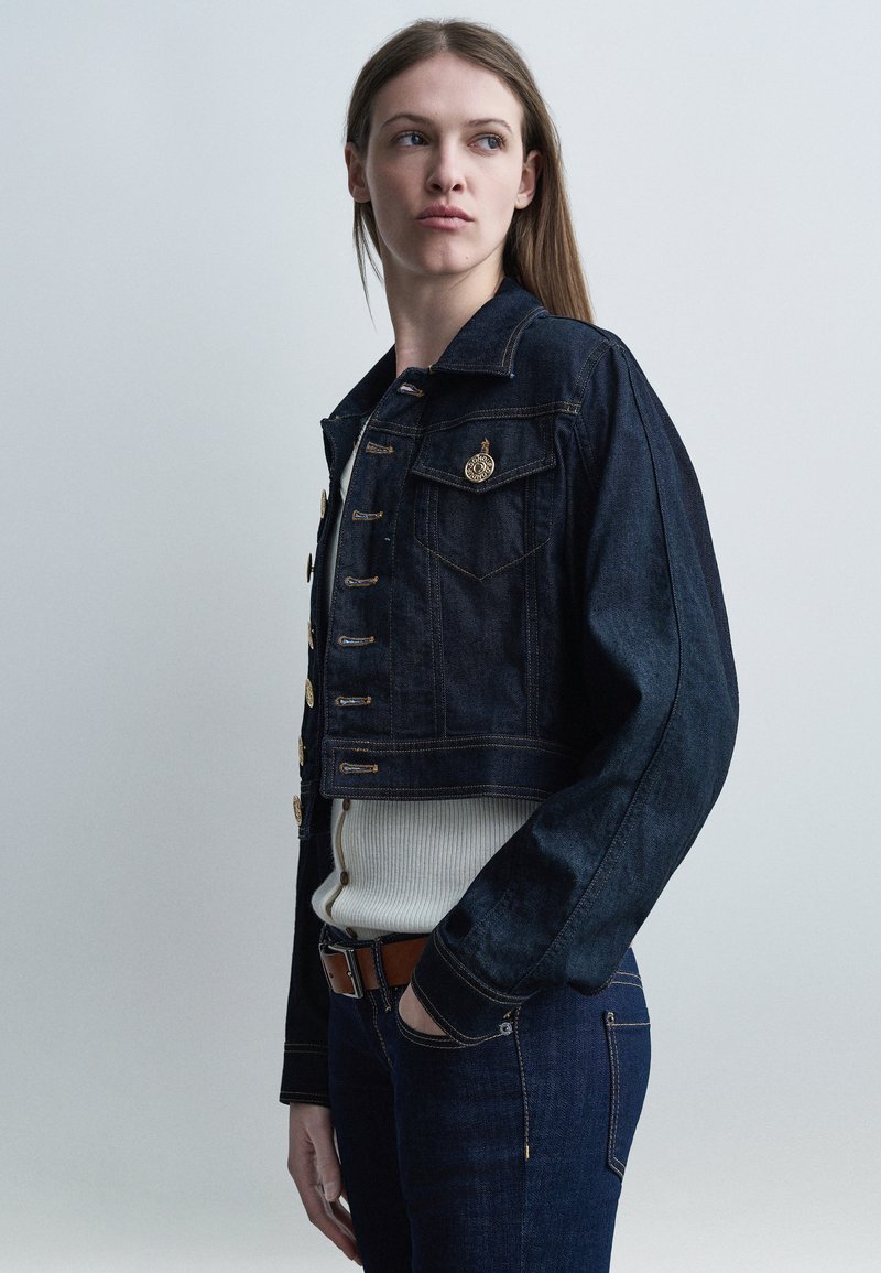 Νεαρή γυναίκα με σκουρόχρωμο τζιν μπουφάν και τζιν παντελόνι, με το χέρι στην τσέπη, κοιτάζοντας πλάγια πάνω σε ένα απλό ανοιχτό γκρι φόντο.