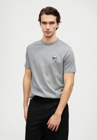 Pilkas medvilnės t-shirt su trumpomis rankovėmis ir apvaliu kaklu, puikuojantis mažu juodu ir baltu piešiniu kairiajame krūtinės šone. Matomos juodos kelnės.