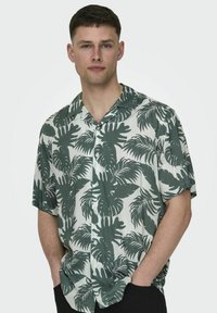 Cămașă cu mâneci scurte, fabricată dintr-un material ușor, cu un model tropical de frunze verzi și albe, având guler cu nasturi și o croială lejeră.