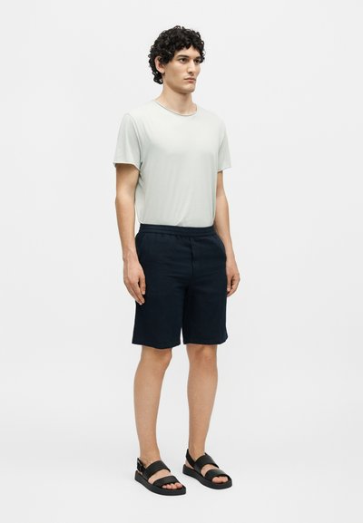 Νέος άνδρας με σγουρά μαλλιά, φορώντας γκρι ανοιχτό t-shirt, σκούρα μπλε σορτς και μαύρα σανδάλια, στέκεται μπροστά σε απλό λευκό φόντο.