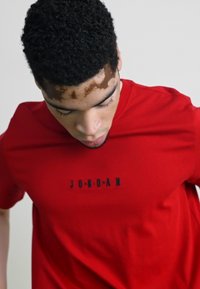 Piros pamut póló, kerek nyakkivágással és egy fekete "JORDAN" logóval a mellkason. Az anyag simának tűnik, enyhe fényével.