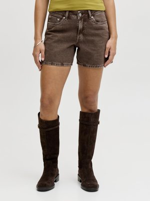 Person iført brune denimshorts, mørkebrune ruskind knæhøje støvler og en gul top mod en ensfarvet baggrund.