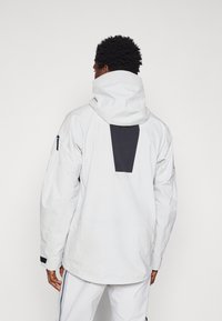 Chaqueta impermeable blanca con un acento negro en la parte posterior, capucha ajustable y bolsillo con cremallera en la manga. Tejido texturizado.