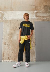 Sort grafisk t-shirt med gul print, sorte træningsbukser, gul jakke bundet om taljen og hvide sneakers med røde detaljer.