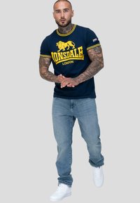 Svalnatý muž s tetováním, oblečený v námořnickém modrém tričku Lonsdale London, světle modrých džínách a bílých teniskách, stojící s rukama spojenýma.