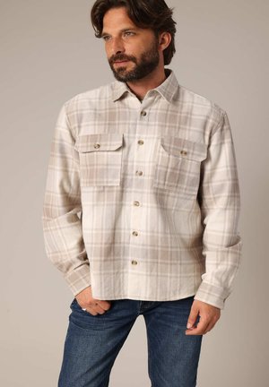 Homme barbu portant une chemise à carreaux beige et blanche avec des poches poitrine et un jean bleu, se tenant devant un fond uni.
