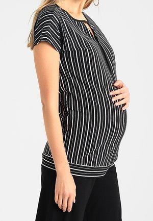 Gravid kvinde holder sin mave, iført en sort og hvid kortærmet top med lodrette striber og sorte bukser, foran en ensfarvet baggrund.