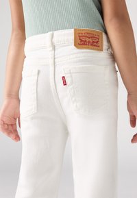 Jean en denim blanc avec poches arrière, présentant une étiquette en cuir marron portant la mention "Levi Strauss & Co." et une petite étiquette de marque rouge sur le côté.