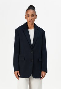 Merino sininen oversized bleiseri, joka on valmistettu sileästä kankaasta. Siinä on kaksi etupainiketta, lovet lapelit ja etutaskut. Päällä vaalea paita.