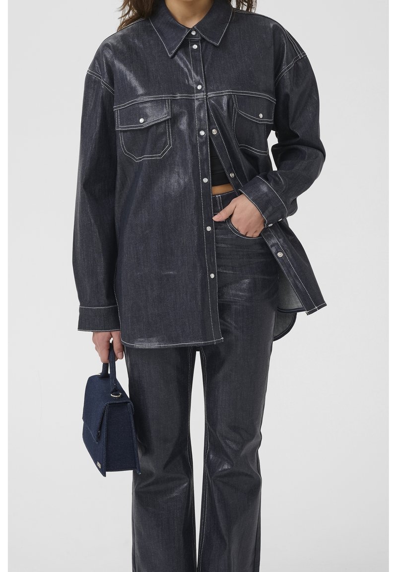 Femme portant une chemise en jean foncé avec des coutures blanches, un pantalon taille haute assorti, tenant un petit sac à main bleu foncé à ses côtés.