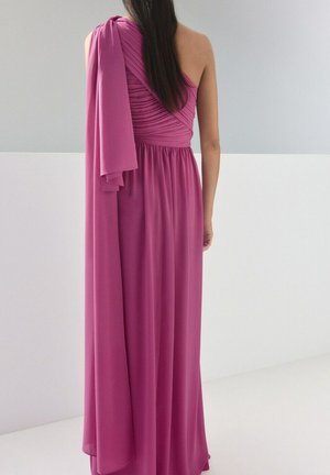 Kobieta o długich ciemnych włosach ubrana w długą, sięgającą podłogi, różową suknię na jedno ramię z drapowanym materiałem na lewym ramieniu, stojąca tyłem.