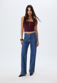 Haut court violet foncé, jean bleu à jambes larges avec poches avant et boutons dorés, porté avec des chaussures noires à bout pointu ; tissu lisse et lignes nettes.