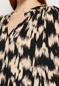 Gros plan d'une femme portant un chemisier beige avec un motif abstrait noir, doté d'un col froncé et en forme de V.