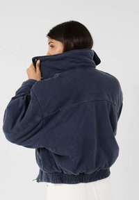 Marineblauw cropped jack met een hoge kraag, elastische tailleband en zachte, gestructureerde stof. Ritssluiting en een ontspannen pasvorm.