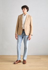 Blazer en lin beige avec deux poches avant, associé à une chemise blanche et un jean bleu clair. Une paire de chaussures en cuir marron complète la tenue.