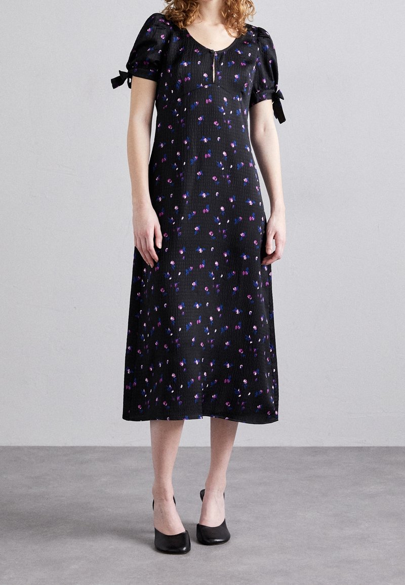 Kvinna som bär en svart midiklänning med puffärmar, små lila och blå blommönster, och svarta klackar med fyrkantig tå, stående på en grå golv.