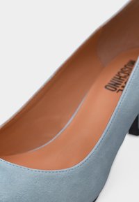 Γυναικείο παπούτσι από απαλή μπλε σουέντ με χαμηλό τακούνι, εσωτερικό από δέρμα καφέ ανοιχτού χρώματος και ραφές στα άκρα, εμφανιζόμενο σε κοντινή λήψη σε λευκό φόντο.